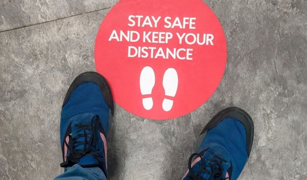 Two feet positioned over a sticker on the ground that reads "Stay Safe, Keep Your Distance," emphasizing the importance of social distancing.